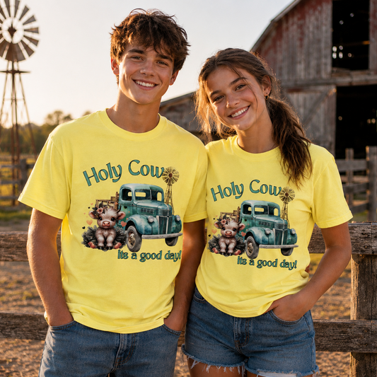 A man and a woman standing outdoors in front of a rustic barn and wooden fence, both wearing bright yellow graphic T-shirts with a vintage truck illustration and the text 'Holy Cow! It's a good day!'.