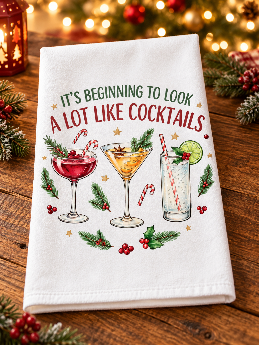 A white kitchen towel with a festive Christmas-themed cocktail print, displayed on a rustic wooden table with holiday lights and pine branches in the background.
