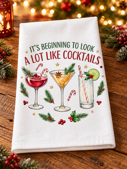 A white kitchen towel with a festive Christmas-themed cocktail print, displayed on a rustic wooden table with holiday lights and pine branches in the background.