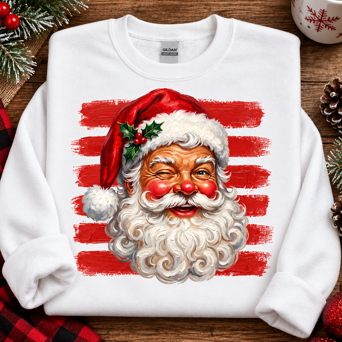 A white Gildan sweatshirt laid flat on a rustic wooden surface, featuring a large graphic of Santa Claus's face over red brushstroke stripes. The scene is styled with holiday props including pine branches, a red plaid fabric, a pinecone, and a ceramic mug.