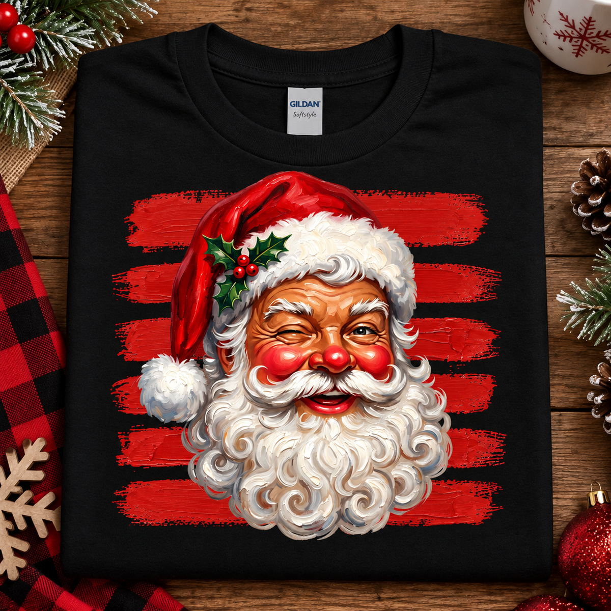 A black Gildan Comfort T-shirt laid flat on a rustic wooden surface, featuring a large graphic of Santa Claus's face with a red hat, white beard, and red striped background. The shirt is styled with holiday decorations including pine branches, a red plaid cloth, a snowflake, pinecones, and red ornaments.