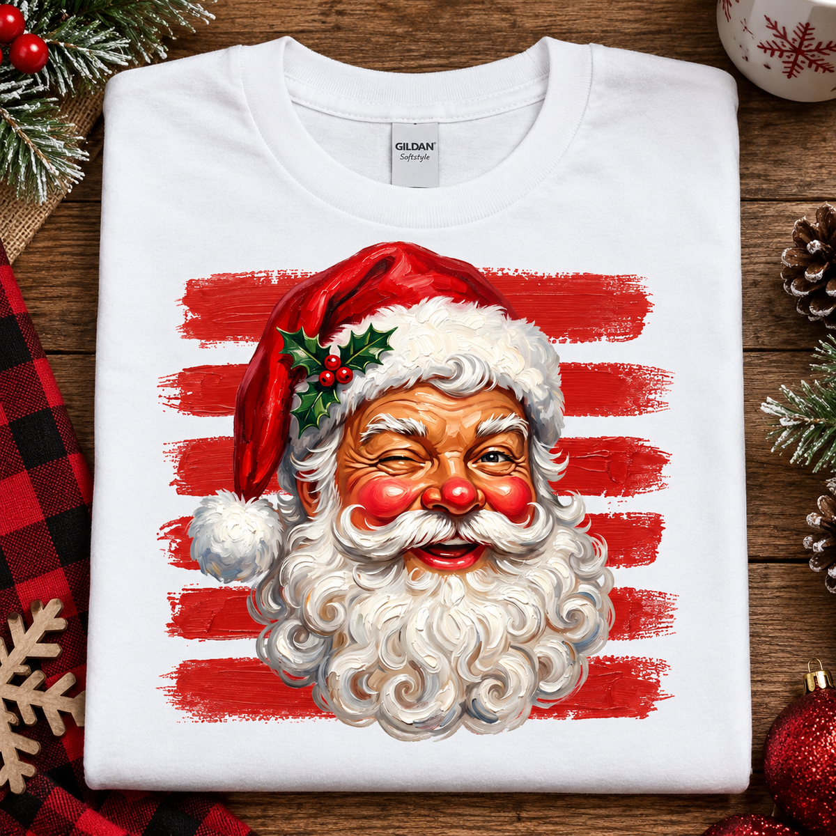 A folded white Gildan Comfort T-shirt with a large graphic of Santa Claus's face on the front, featuring red and white horizontal brushstroke stripes in the background and holiday greenery accents. The shirt is styled in a flat-lay on a rustic wooden surface with Christmas decorations.