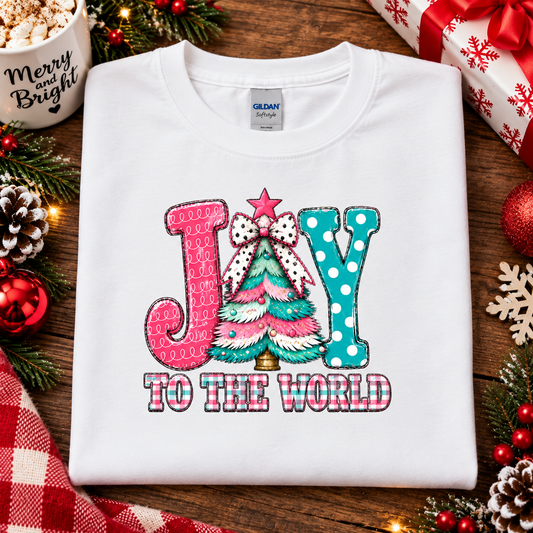 A folded white Gildan t-shirt with a colorful Christmas graphic reading 'JOY TO THE WORLD' in pink and teal, surrounded by holiday decorations like pine branches, ornaments, a gift box, and a mug on a wooden surface.