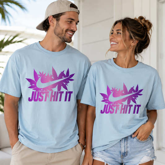 A man and a woman wearing matching light blue t-shirts with a purple and pink graphic of a Nike swoosh layered over a palm leaf and the text 'JUST HIT IT'. The man is wearing a beige baseball cap backwards, and they are standing on a sunny porch with a white house and palm trees in the background.
