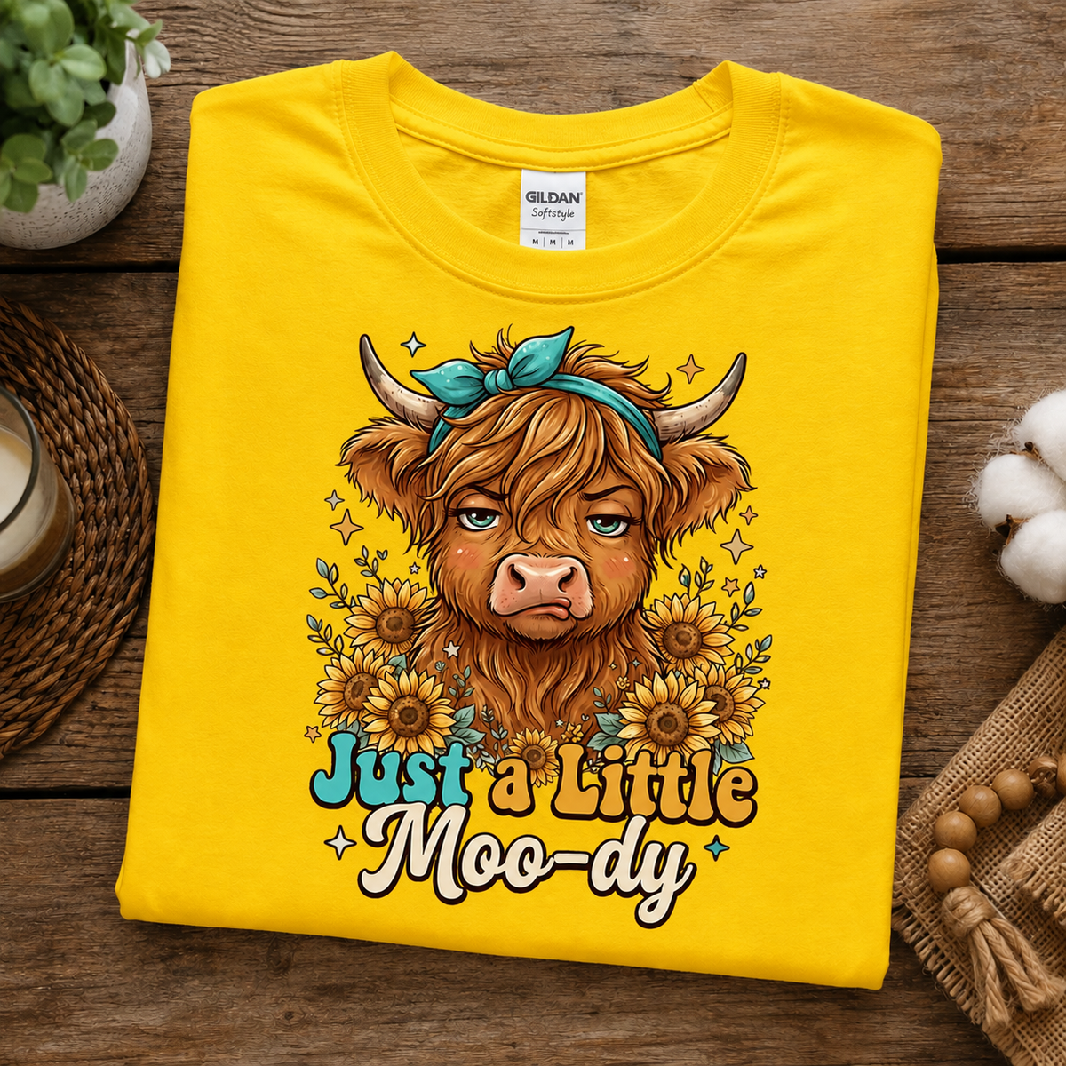 A folded bright yellow Gildan Comfort T-shirt is displayed on a rustic wooden surface. The shirt features a graphic of a shaggy Highland cow with a teal bow, surrounded by sunflowers, with the text 'Just a Little Moo-dy' written below in a playful teal and brown font.