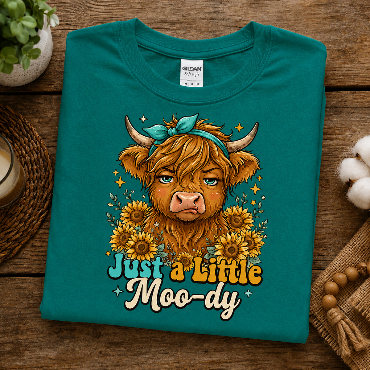 A folded jade dome Gildan t-shirt with a graphic of a sad Highland cow surrounded by sunflowers and the text 'Just a Little Moo-dy', displayed on a rustic wooden background with decorative props.