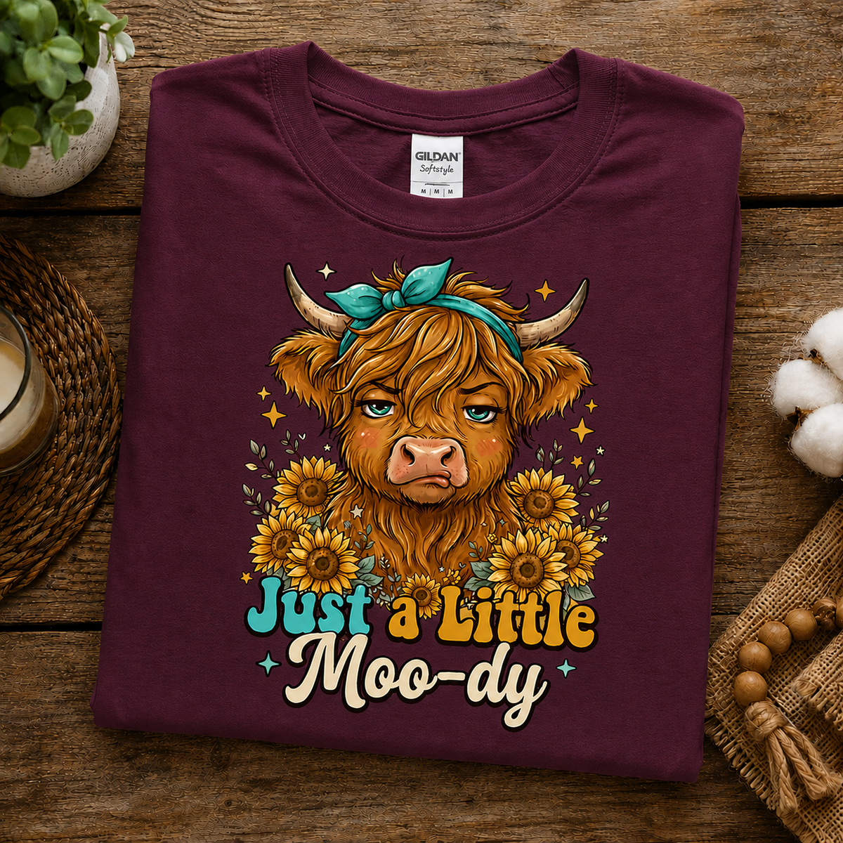 A folded maroon Gildan t-shirt with a graphic of a Scottish Highland cow surrounded by sunflowers and the text 'Just a Little Moo-dy', displayed on a rustic wooden background with decorative props.