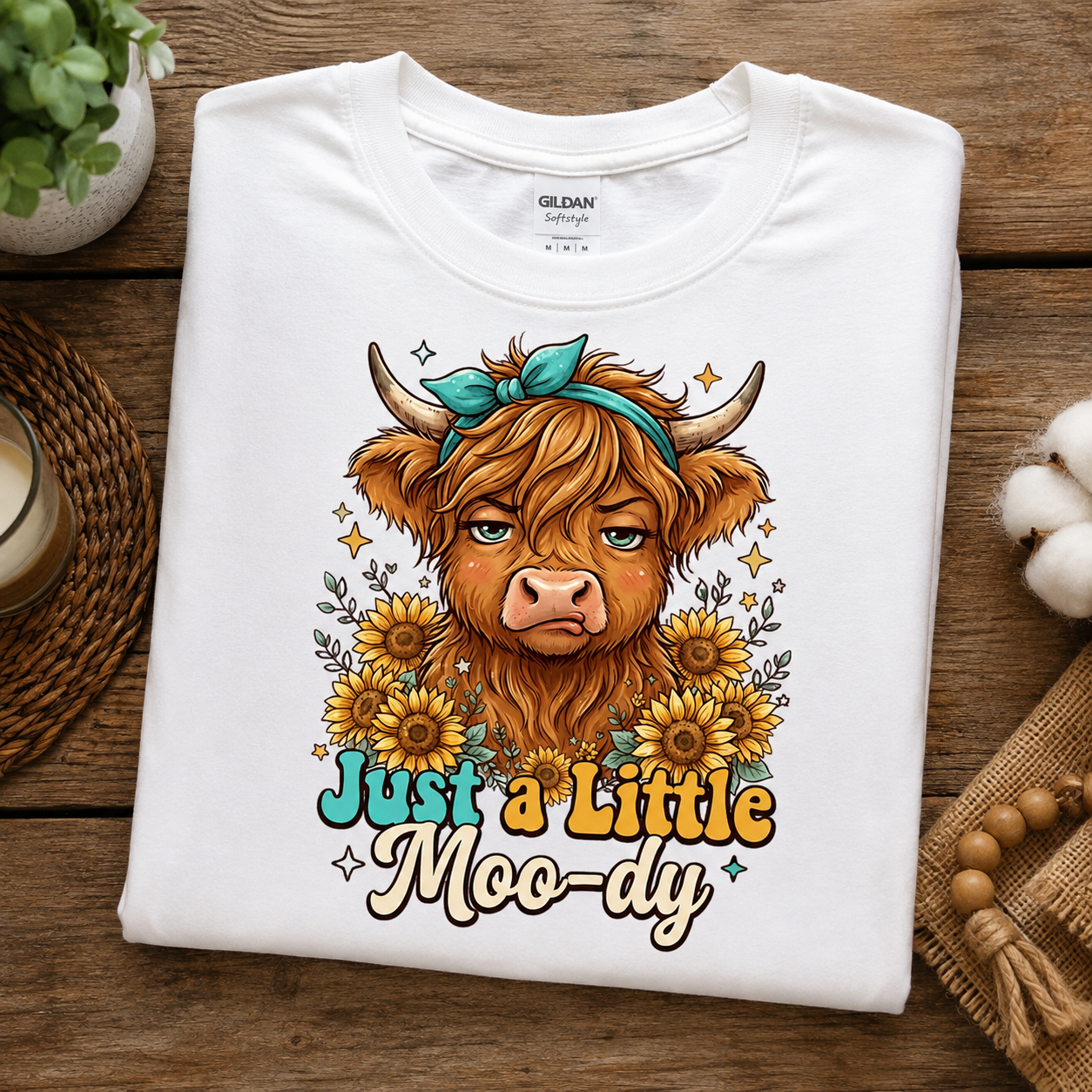 A folded white Gildan Softstyle T-shirt with a colorful graphic of a highland cow surrounded by sunflowers and the text 'Just a Little Moo-dy', displayed on a rustic wooden surface with decorative props.