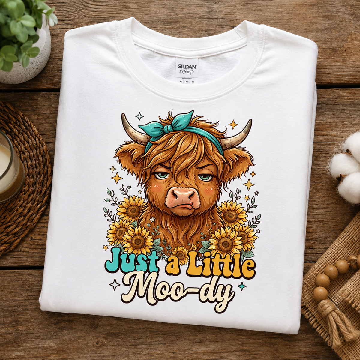 A folded white Gildan Softstyle T-shirt with a colorful graphic of a highland cow surrounded by sunflowers and the text 'Just a Little Moo-dy', displayed on a rustic wooden surface with decorative props.
