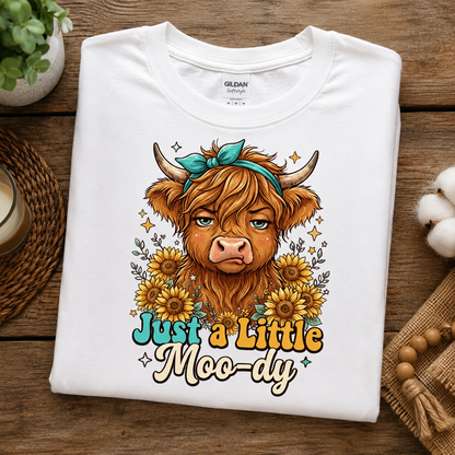 A folded white Gildan Softstyle T-shirt with a colorful graphic of a highland cow surrounded by sunflowers and the text 'Just a Little Moo-dy', displayed on a rustic wooden surface with decorative props.