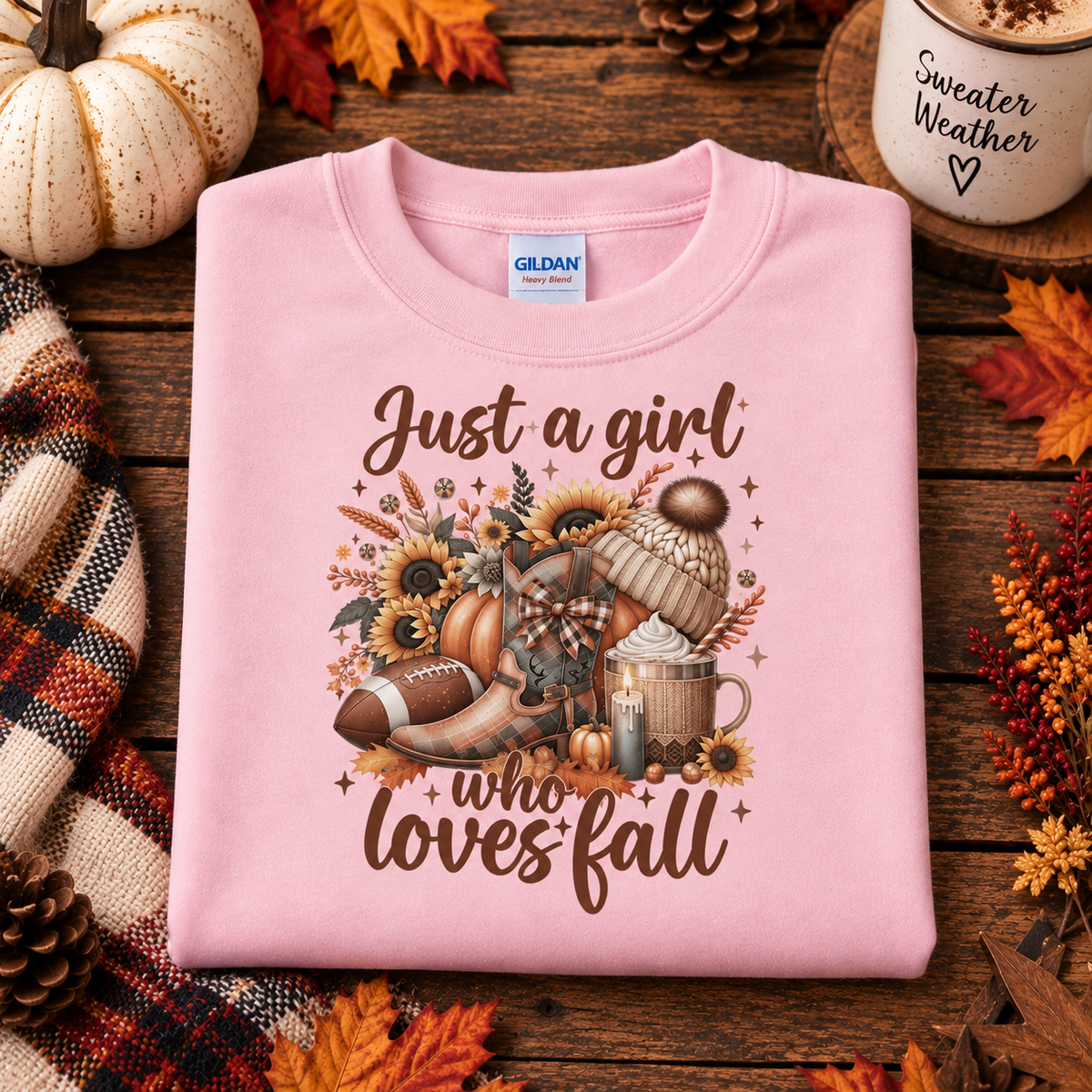 A folded light pink crewneck sweatshirt with a fall-themed graphic reading 'Just a girl who loves fall' in brown script. The graphic includes autumn elements like sunflowers, a football, a mug, and pumpkins.