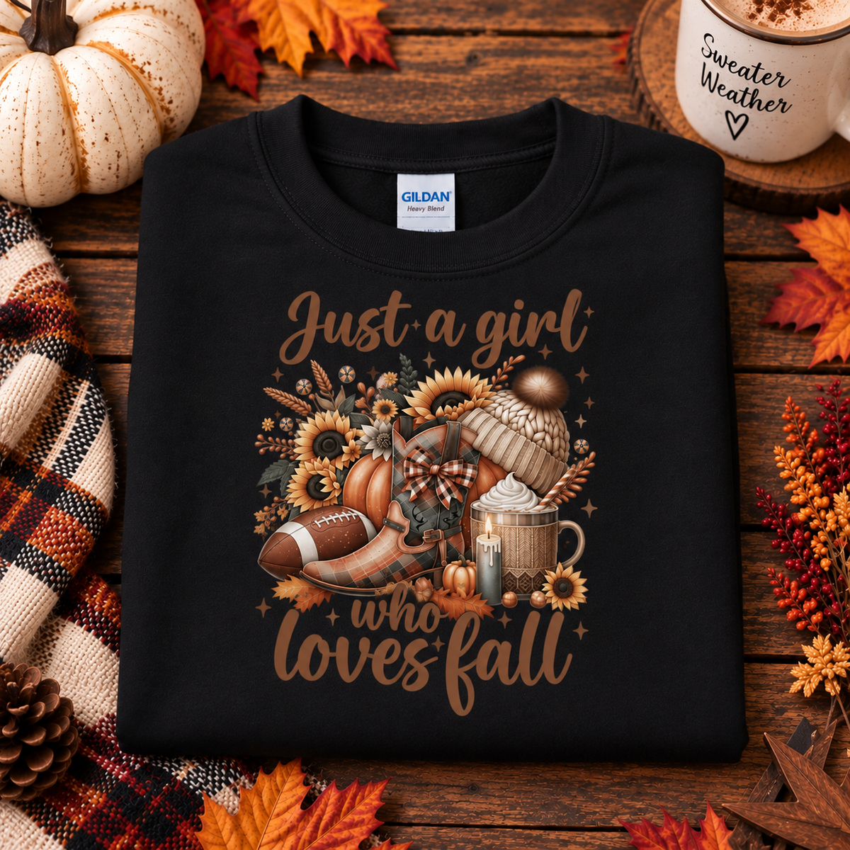 A black Gildan sweatshirt laid flat on a rustic wooden surface, surrounded by autumn props including pumpkins, fall leaves, a plaid scarf, and a mug that reads 'Sweater Weather'. The sweatshirt features a large graphic with the phrase 'Just a girl who loves fall' in brown script, decorated with sunflowers, a football, a pumpkin, a mug with whipped cream, and a beanie.
