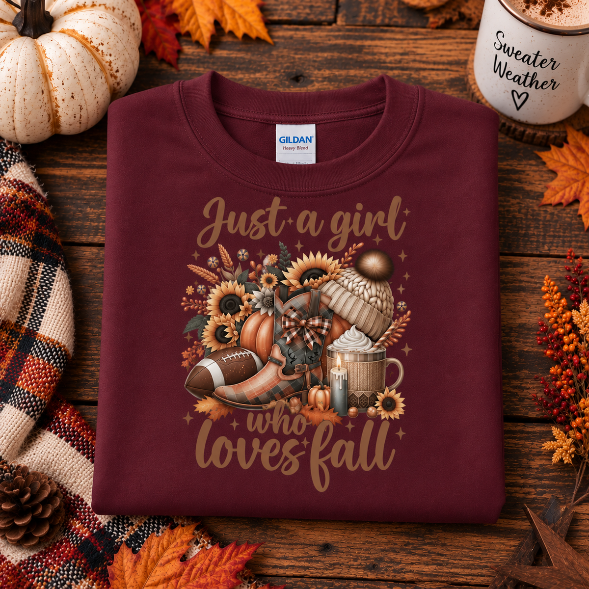 A folded maroon Gildan t-shirt with a 'Just a girl who loves fall' graphic featuring autumn elements, styled on a rustic wooden background with pumpkins, a plaid blanket, and a coffee mug.