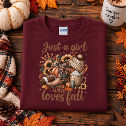 A folded maroon Gildan t-shirt with a 'Just a girl who loves fall' graphic featuring autumn elements, styled on a rustic wooden background with pumpkins, a plaid blanket, and a coffee mug.