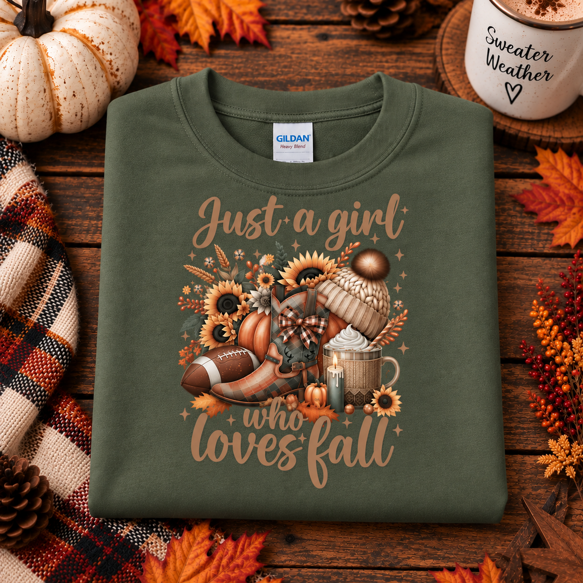 An military green Gildan t-shirt with a 'Just a girl who loves fall' graphic print featuring autumn elements like a football, pumpkin, and mug, styled in a flat-lay on a wooden surface with fall decorations.