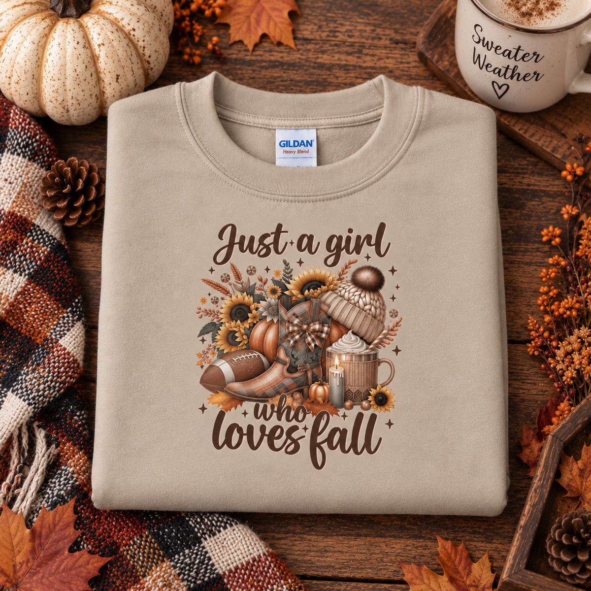 A folded sand crewneck sweatshirt with a graphic print reading 'Just a girl who loves fall' surrounded by autumn-themed illustrations, styled on a wooden surface with a pumpkin, plaid scarf, and a mug that says 'Sweater Weather'.