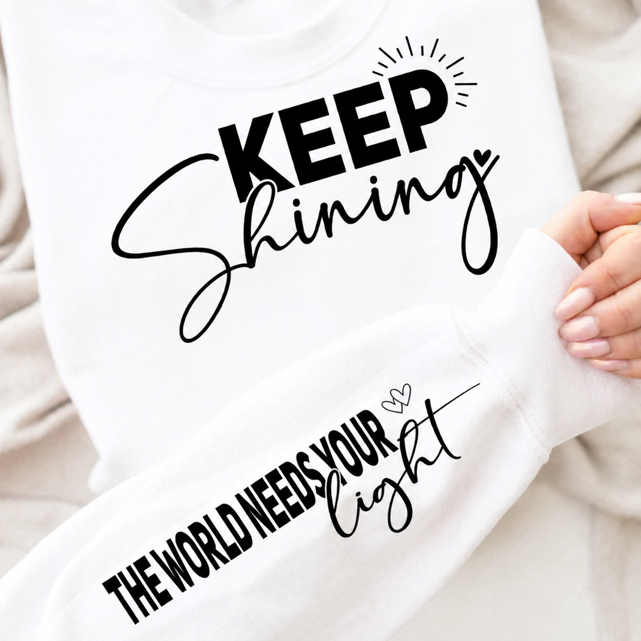 White sweatshirt with motivational text 'Keep Shining, The World Needs Your Light' held by a person.