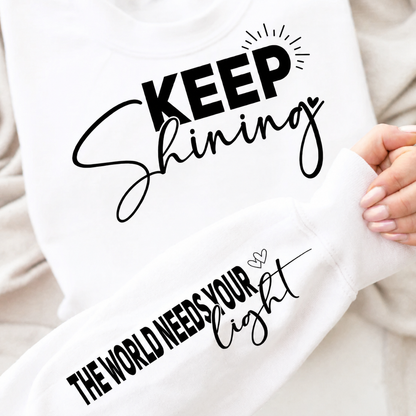 White sweatshirt with motivational text 'Keep Shining, The World Needs Your Light' held by a person.