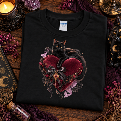 A folded black Gildan t-shirt with a graphic of a black cat resting on a large red heart with a black ribbon and purple flowers, styled on a rustic wooden table with candles, crystals, and a moon figurine.