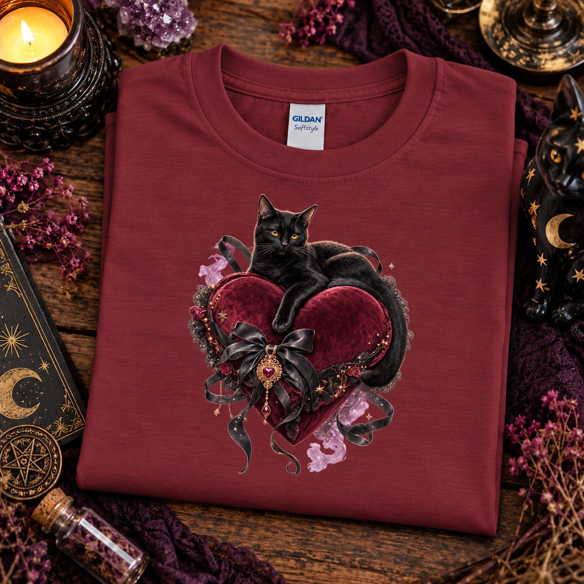 A folded Gildan Spectrum t-shirt in a garnet red, featuring a graphic print of a black cat resting on a dark red heart with a black ribbon and small pink flowers. The shirt is styled in a cozy, mystical flat-lay setting with a lit candle, purple crystals, a starry night lantern, and a book on a rustic wooden table.