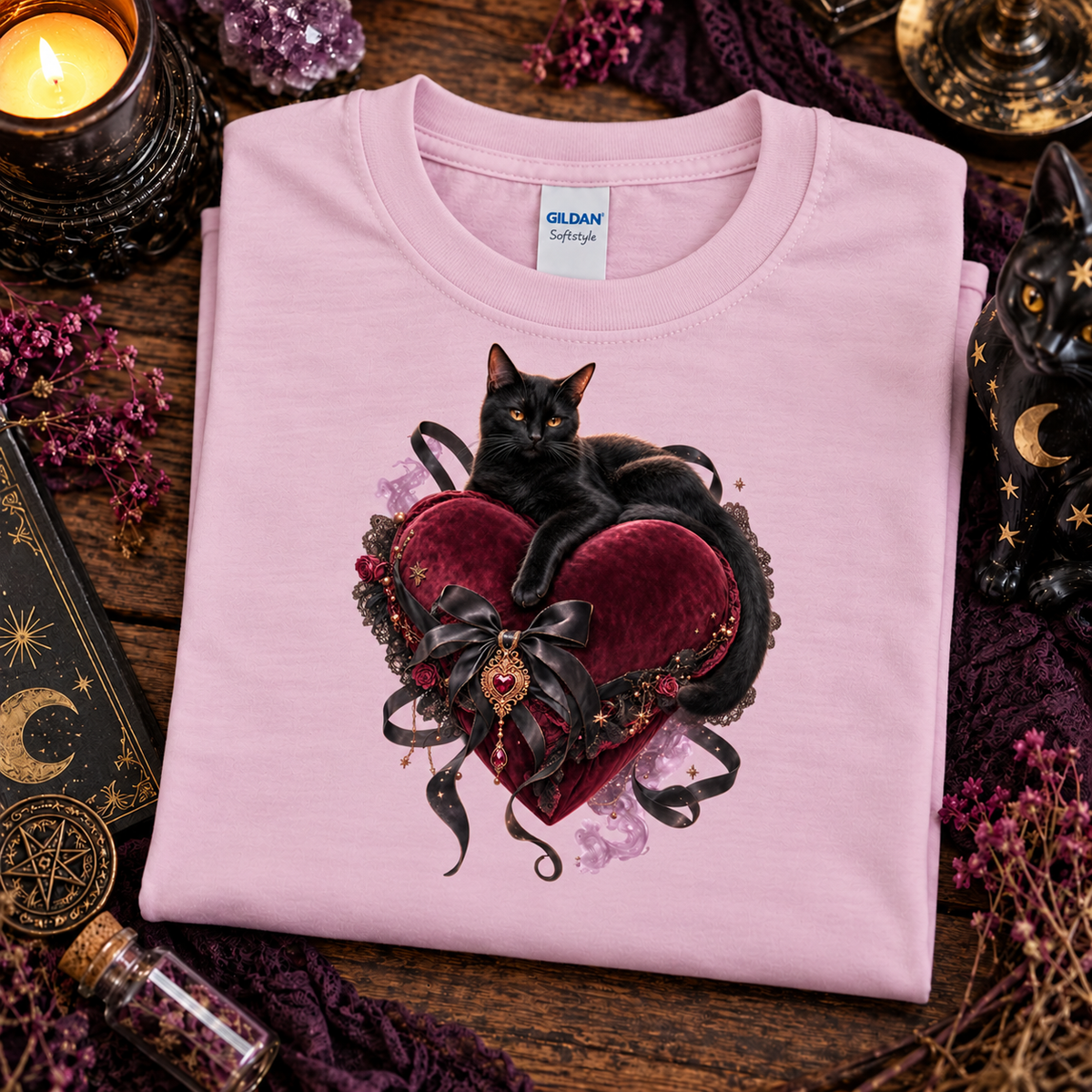 A folded light pink Gildan Softstyle T-shirt with a graphic of a black cat resting on a dark red velvet heart, surrounded by mystical props like a candle, crystals, and a black cat figurine on a wooden surface.