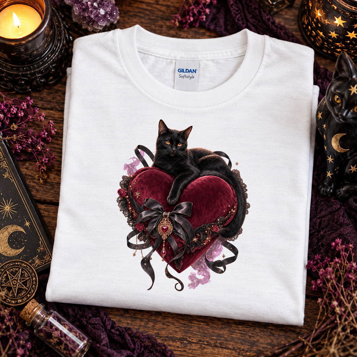 A folded white Gildan t-shirt with a graphic of a black cat resting on a dark red velvet heart with black lace and ribbon details, styled on a wooden surface with occult-themed props like candles, a starry orb, and a cat figurine.