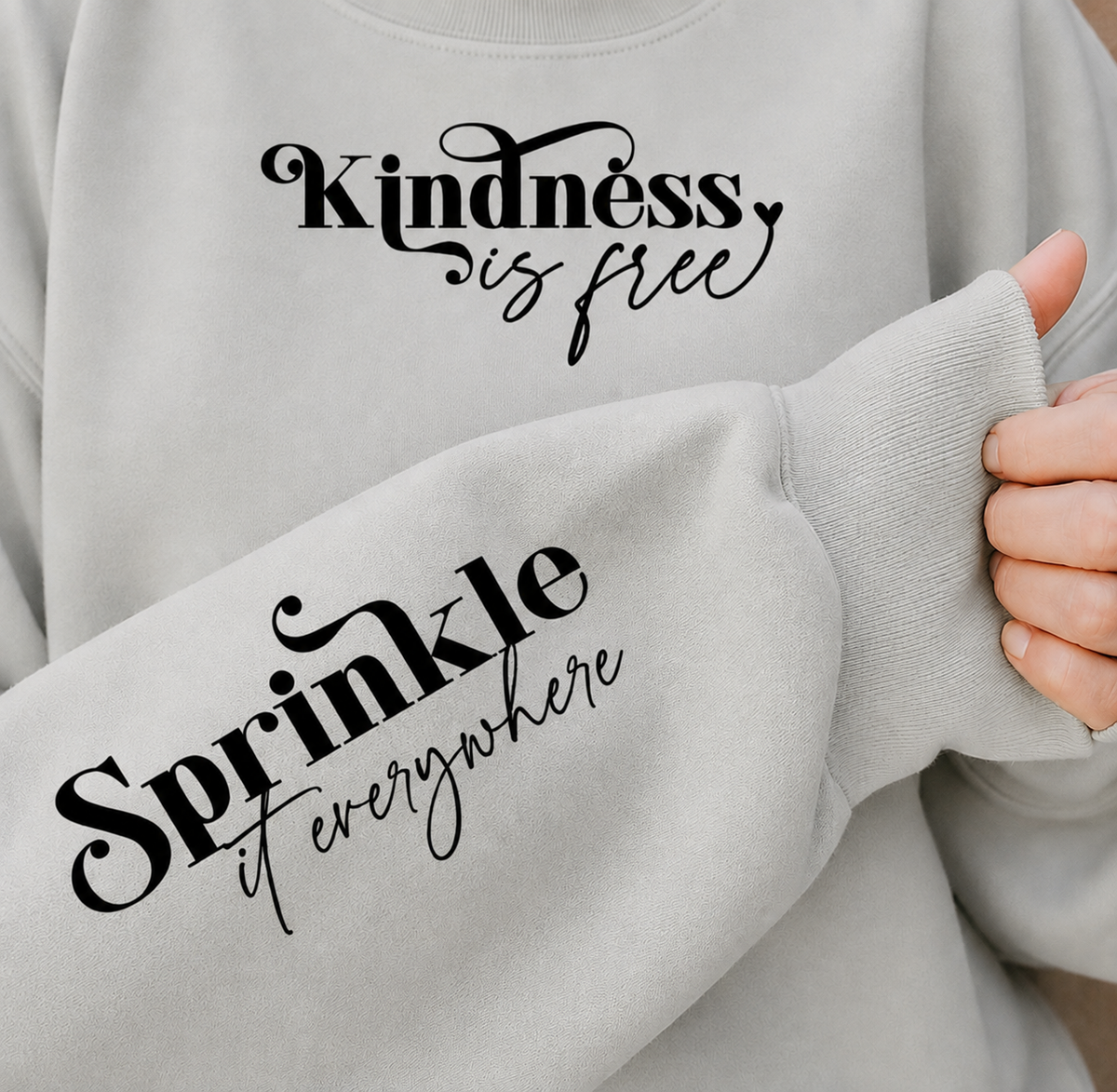 A close-up of a light gray crewneck sweatshirt with black printed text. The chest reads 'Kindness is free' and the sleeve reads 'Sprinkle it everywhere'.