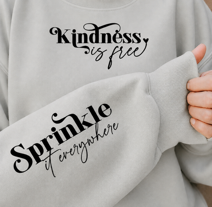 A close-up of a light gray crewneck sweatshirt with black printed text. The chest reads 'Kindness is free' and the sleeve reads 'Sprinkle it everywhere'.
