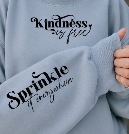 A close-up of a light blue sweatshirt worn by a person, showing the front chest text 'Kindness is free' and the sleeve text 'Sprinkle it everywhere' in black script. A hand is holding the ribbed cuff of the sleeve.