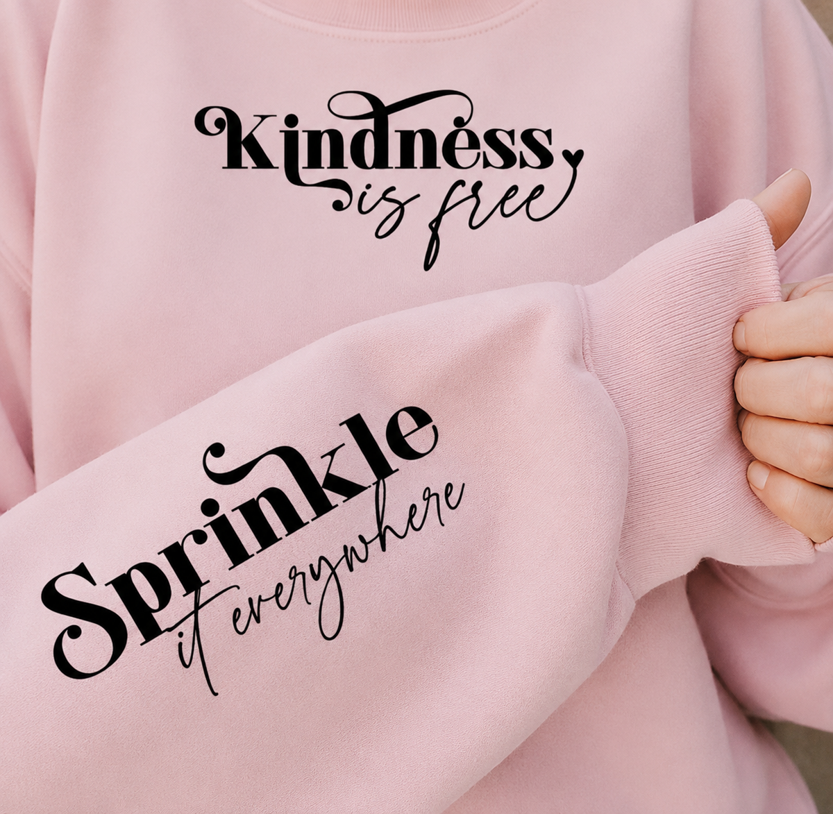 A close-up of a light pink sweatshirt with black script text reading 'Kindness is free' on the chest and 'Sprinkle it everywhere' on the sleeve, worn by a person whose hand is visible holding the cuff.