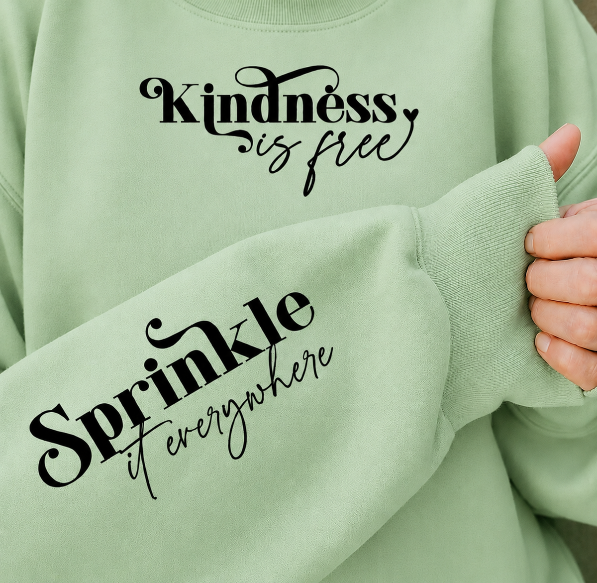 A close-up of a mint green crewneck sweatshirt with black script text on the chest reading 'Kindness is free' and on the sleeve reading 'Sprinkle it everywhere'. A person's hand is holding up the sleeve cuff to show the fabric texture.