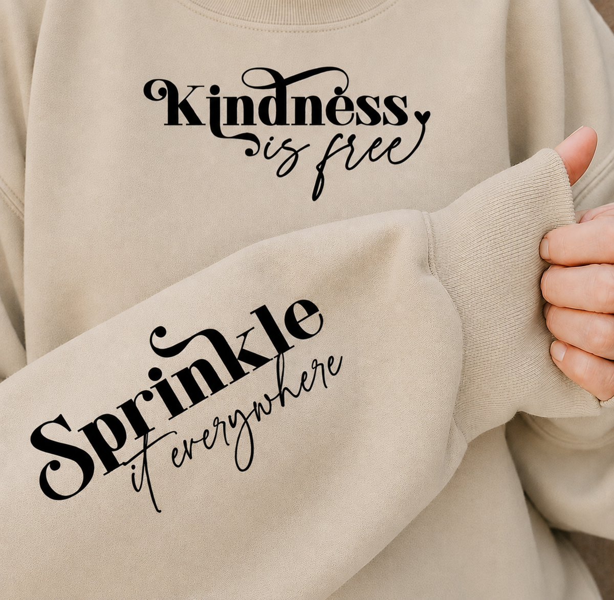 A close-up of a sand colored sweatshirt with black script text reading 'Kindness is free' on the chest and 'Sprinkle it everywhere' on the sleeve, worn by a person whose hand is visible holding the cuff.
