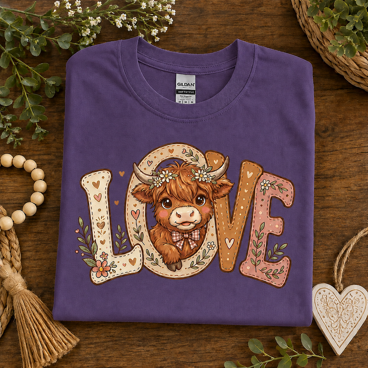 A folded heather purple t-shirt with a 'LOVE' graphic featuring a cute brown Highland cow, styled on a rustic wooden background with decorative greenery and beads.