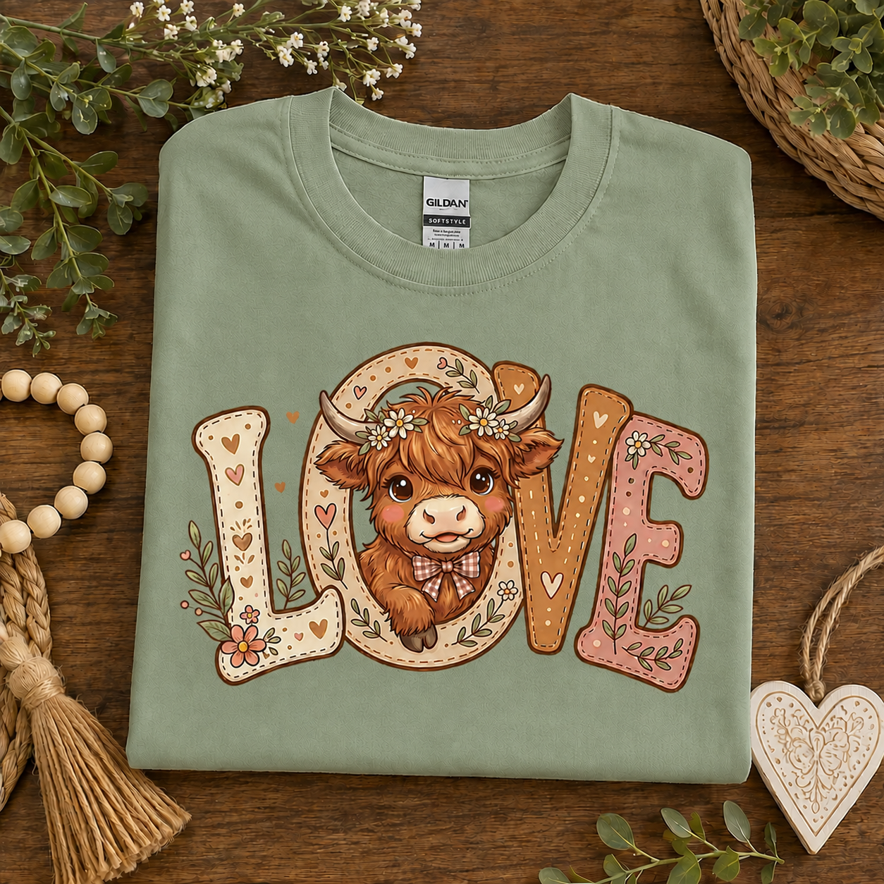 A folded sage green t-shirt with a 'LOVE' cow graphic, displayed on a rustic wooden background with decorative eucalyptus leaves, beads, and a heart-shaped ornament.
