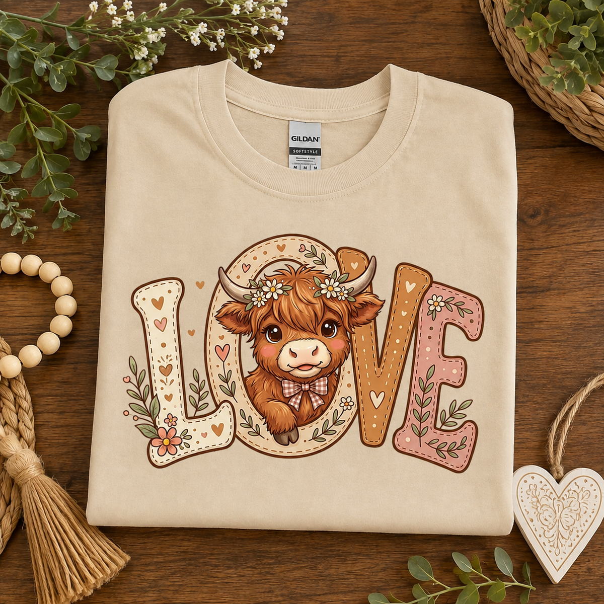 A folded sand colored Gildan t-shirt with a 'LOVE' graphic featuring a brown cow illustration, styled on a dark wooden background with decorative greenery and a heart-shaped ornament.