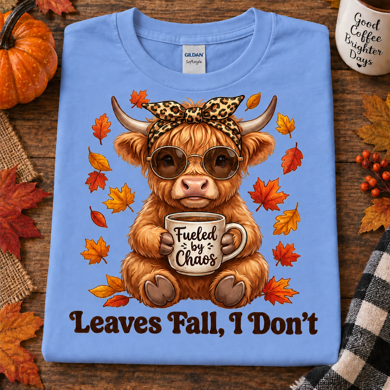 A folded carolina blue Gildan Softstyle t-shirt is displayed on a rustic wooden background with autumn props. The shirt features a graphic of a fluffy Highland cow wearing sunglasses and a leopard-print bow, holding a mug that says 'Fueled by Chaos', surrounded by falling leaves, with the text 'Leaves Fall, I Don't' printed below.