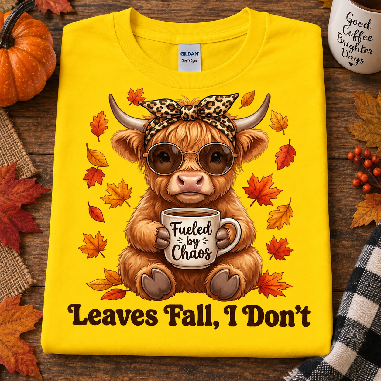 A folded daisy colored graphic T-shirt is displayed on a rustic wooden surface with autumn props. The shirt features a cartoon Highland cow wearing sunglasses and a leopard-print bow, holding a mug that says 'Fueled by Chaos,' surrounded by falling leaves, with the text 'Leaves Fall, I Don't' below.
