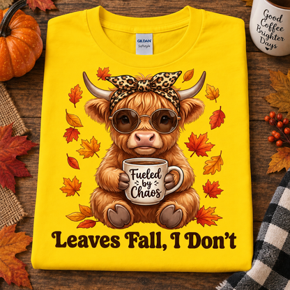 A folded daisy colored graphic T-shirt is displayed on a rustic wooden surface with autumn props. The shirt features a cartoon Highland cow wearing sunglasses and a leopard-print bow, holding a mug that says 'Fueled by Chaos,' surrounded by falling leaves, with the text 'Leaves Fall, I Don't' below.