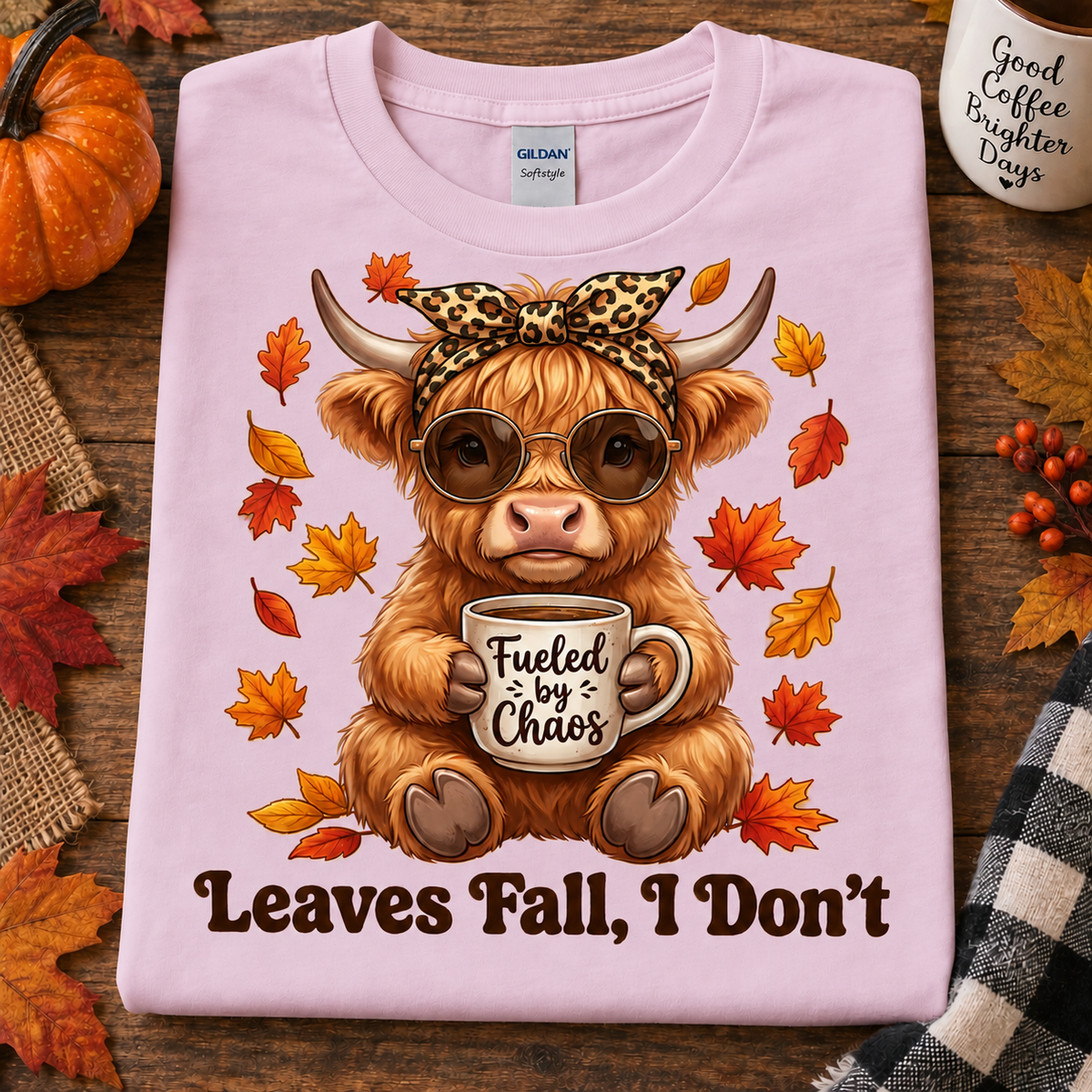 A folded light pink Gildan Softstyle t-shirt is displayed on a rustic wooden surface with autumn props. The shirt features a graphic of a fluffy brown highland cow wearing sunglasses and a leopard-print bow headband, holding a mug that says 'Fueled by Chaos'.