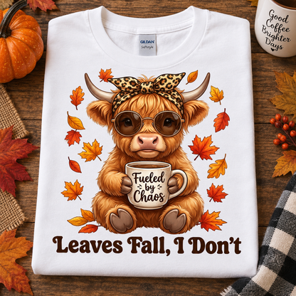 A folded white Gildan Softstyle T-shirt is displayed on a rustic wooden surface with autumn props. The shirt features a graphic of a fluffy tan Highland cow wearing round sunglasses and a leopard-print bow headband, holding a mug that says 'Fueled by Chaos.