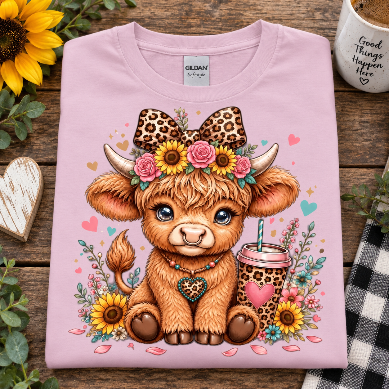 A folded light pink Gildan t-shirt with a colorful graphic of a cute brown highland cow, displayed on a rustic wooden background with decorative props like sunflowers and a mug.