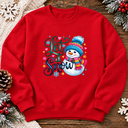 A red crewneck sweatshirt featuring a festive graphic with the text 'Let it Snow' and a cartoon snowman wearing a blue and pink hat and scarf. The sweatshirt is displayed in a flat-lay setting on a rustic wooden surface, surrounded by holiday decorations including pinecones, snow branches, and a knitted blanket.