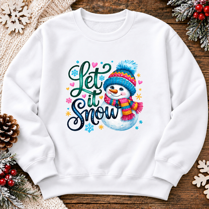 A white crewneck sweatshirt with a colorful 'Let it Snow' graphic featuring a cartoon snowman wearing a blue and pink hat and scarf, surrounded by festive holiday decorations on a wooden surface.