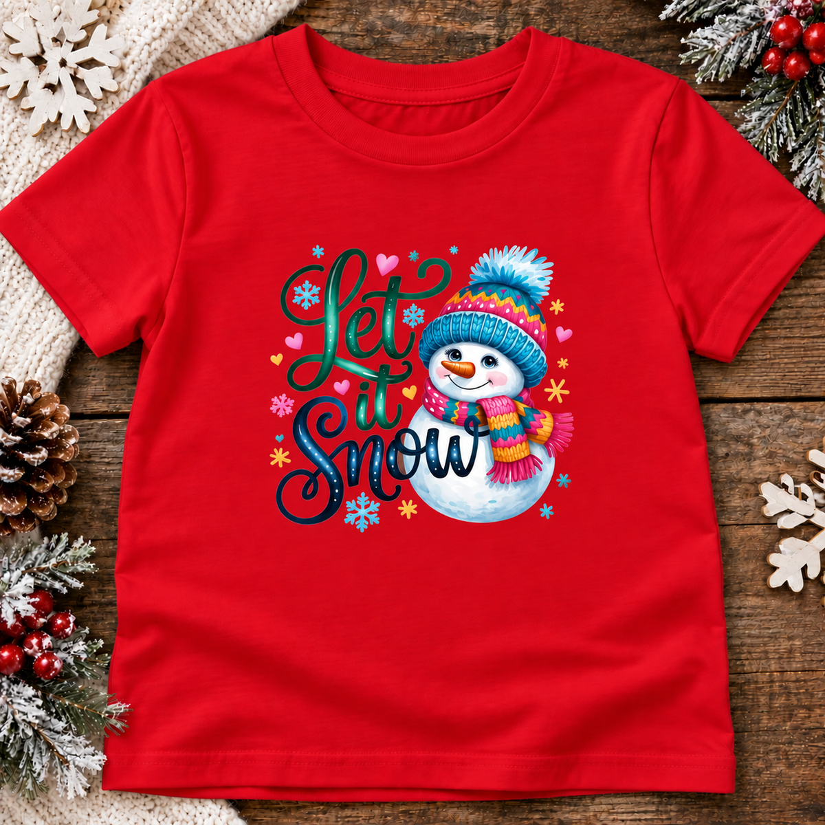 A red short-sleeve kids' t-shirt with a colorful 'Let it Snow' graphic featuring a cartoon snowman wearing a blue hat and scarf, surrounded by festive snowflakes and hearts. The shirt is displayed flat on a rustic wooden surface decorated with pinecones, snow-dusted pine branches, and white ornaments.