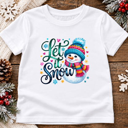 A white short-sleeve t-shirt with a colorful 'Let it Snow' graphic featuring a cartoon snowman wearing a blue and pink hat and scarf, displayed on a rustic wooden background with holiday decorations.