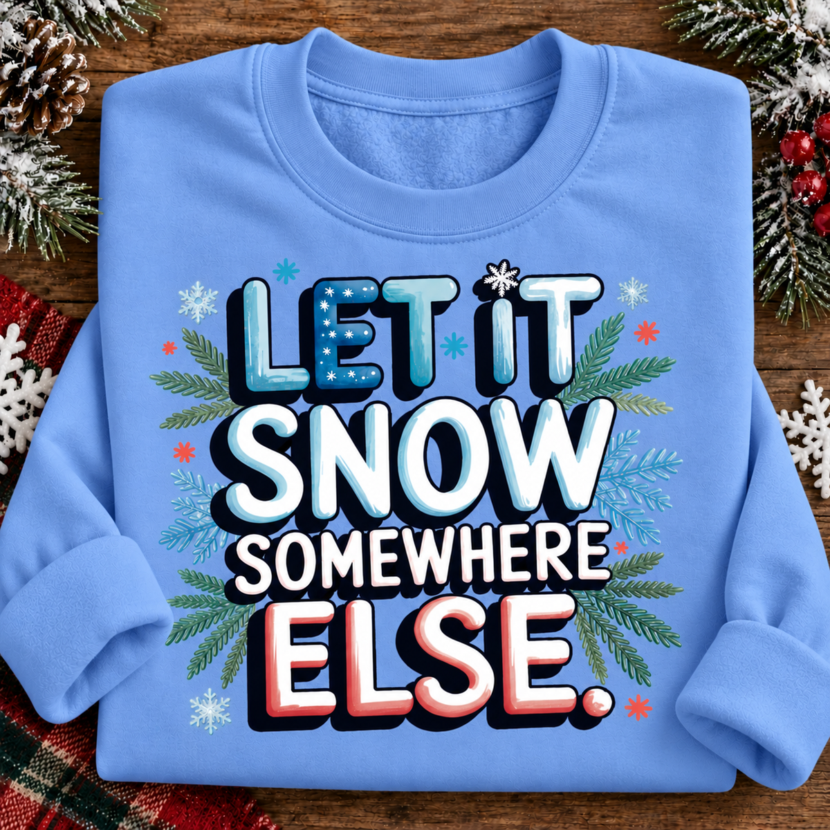 A folded carolina blue crewneck sweatshirt on a rustic wooden background, decorated with pine branches, snowflakes, and red berries. The front features a bold graphic text that reads 'LET IT SNOW SOMEWHERE ELSE.