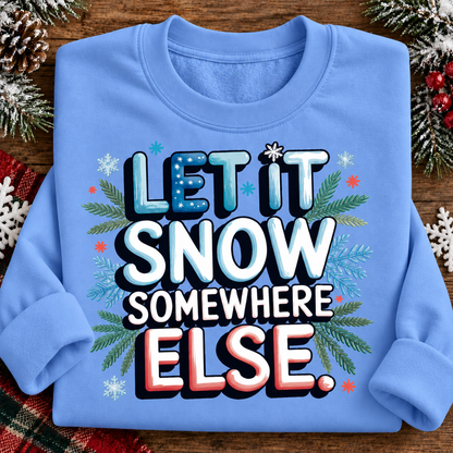 A folded carolina blue crewneck sweatshirt on a rustic wooden background, decorated with pine branches, snowflakes, and red berries. The front features a bold graphic text that reads 'LET IT SNOW SOMEWHERE ELSE.