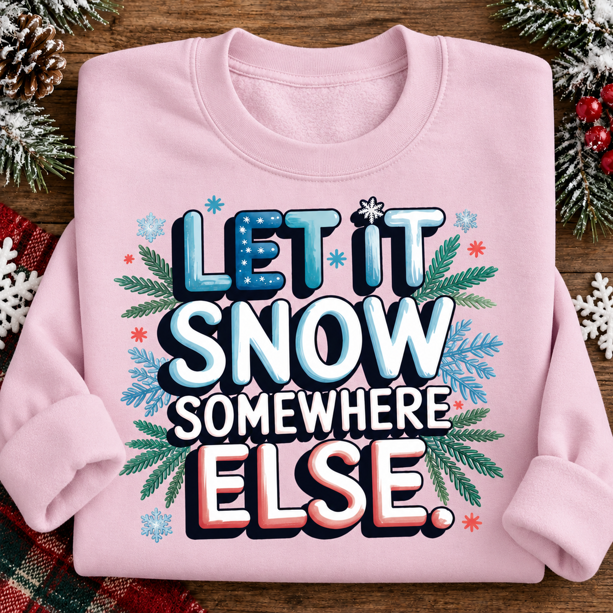 A folded light pink crewneck sweatshirt with a festive graphic reading 'LET IT SNOW SOMEWHERE ELSE.' in blue, white, and red lettering, surrounded by snowflakes, pine branches, and small red stars, displayed on a wooden surface with holiday decorations.