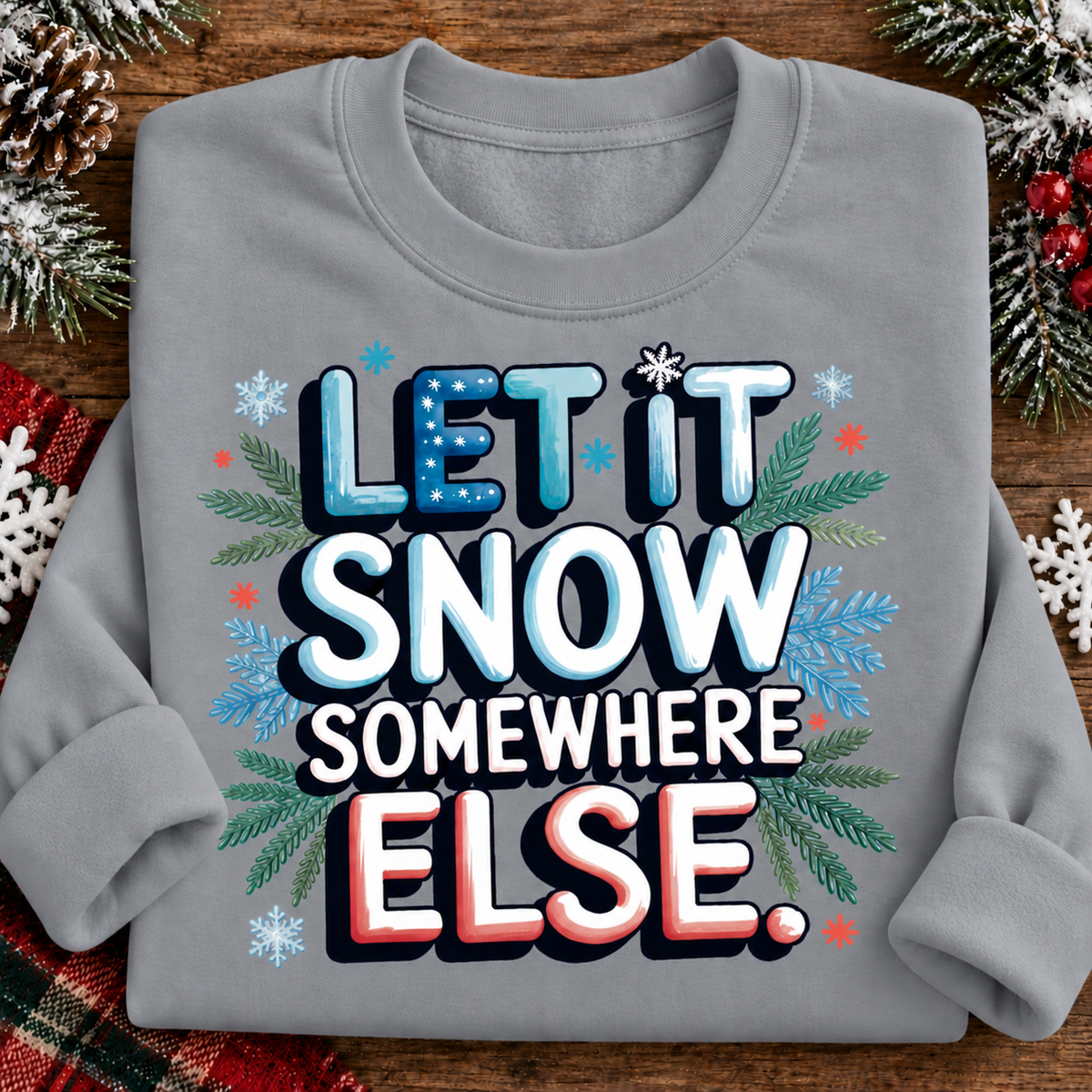 A folded sport gray crewneck sweatshirt with a festive graphic that reads 'LET IT SNOW SOMEWHERE ELSE.' in blue, white, and red lettering, surrounded by snowflakes, pine branches, and small red stars, displayed on a wooden surface with holiday decorations.