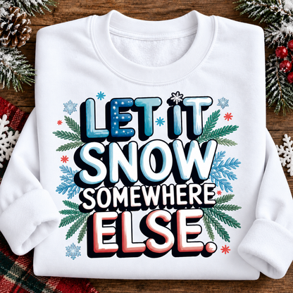 A folded white long-sleeve sweatshirt with a festive graphic reading 'LET IT SNOW SOMEWHERE ELSE' surrounded by snowflakes, pine branches, and red berries, displayed on a wooden surface with holiday decorations.
